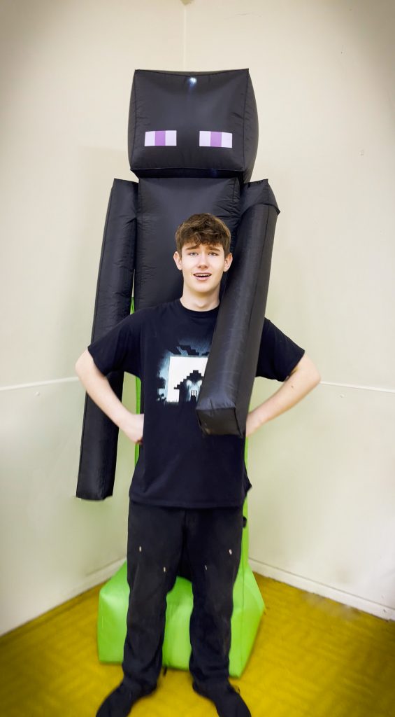 Entertainer wearing a Minecraft T‑shirt poses under the towering Enderman at a hall in Farnborough – size reference for parents.