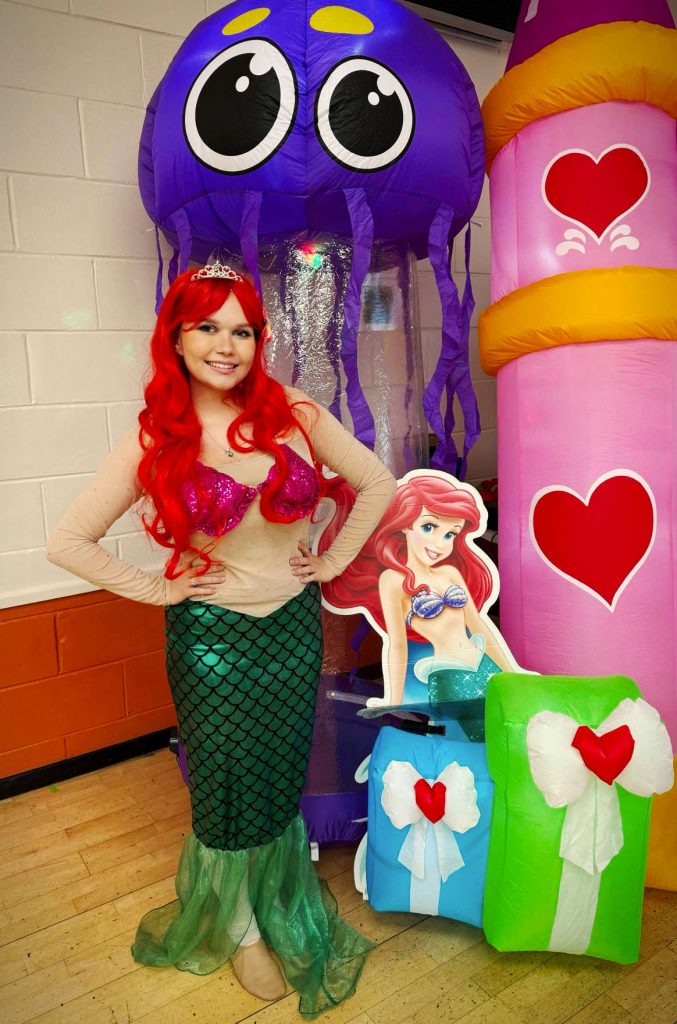 Happy mermaid children's party entertainer with red hair and sparkly seashell top standing beside inflatable jellyfish and castle at Mayford Village Hall near Woking
