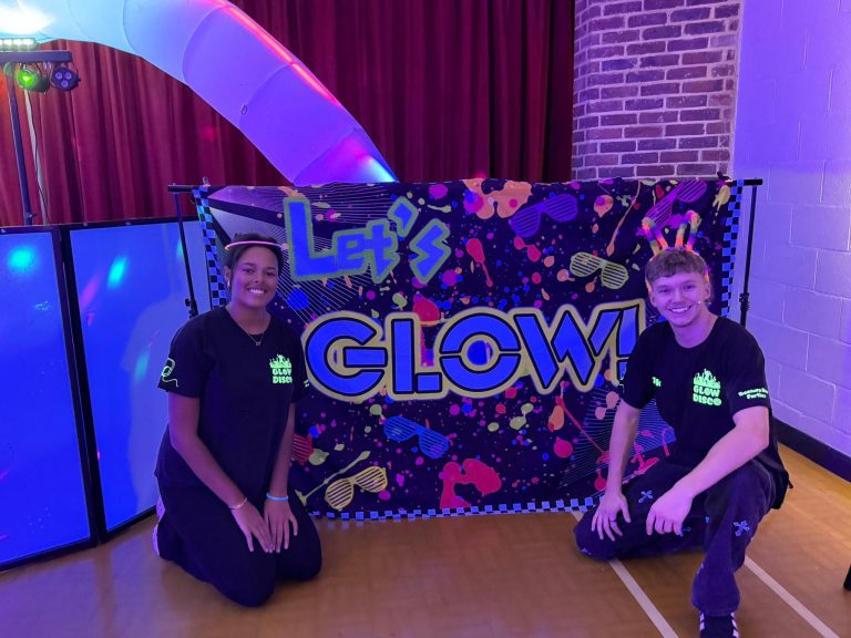 Two glow disco entertainers kneeling beside a Let's Glow neon backdrop