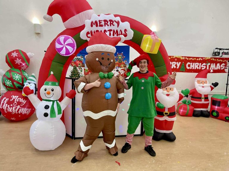 Gingerbread man mascot and an elf under a Merry Christmas arch with Santa inflatables