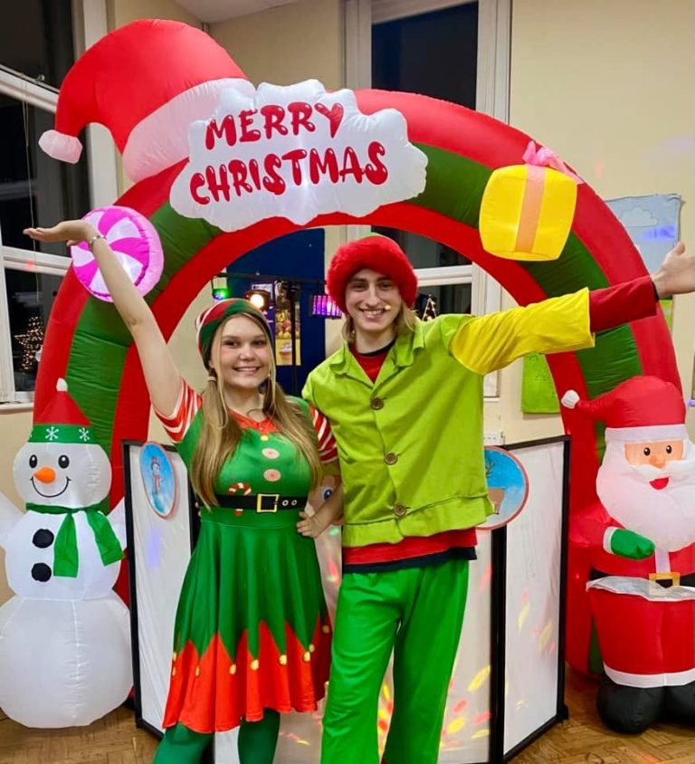 Two elves under a Merry Christmas arch with snowman and Santa inflatables