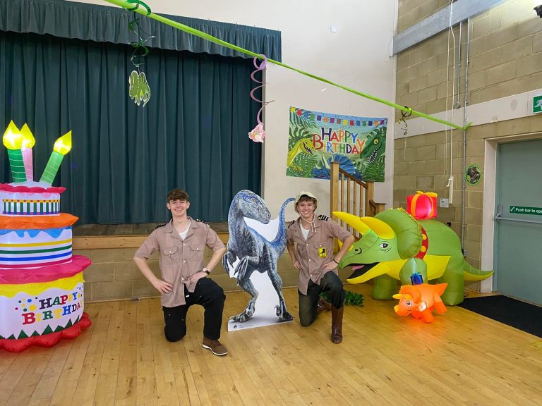 Two explorer entertainers kneeling either side of a raptor cut-out with a triceratops inflatable