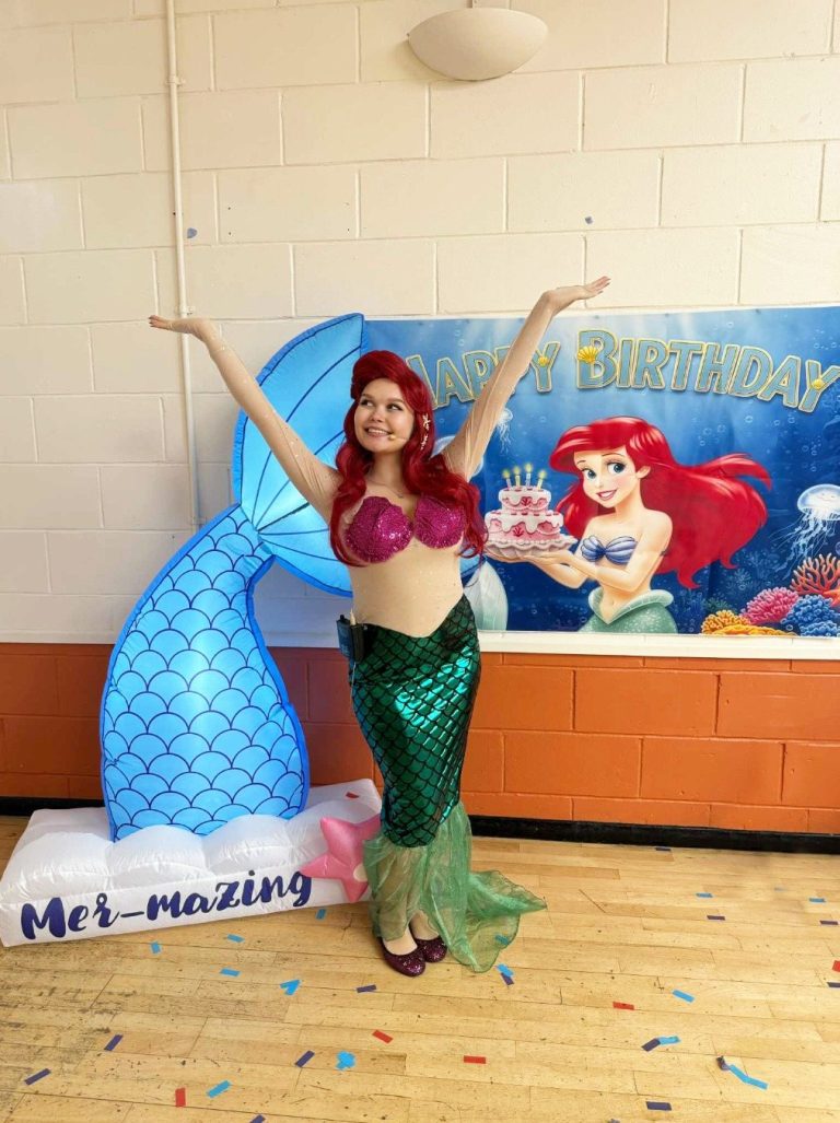Ariel entertainer with arms raised beside a mermaid tail inflatable and Little Mermaid backdrop