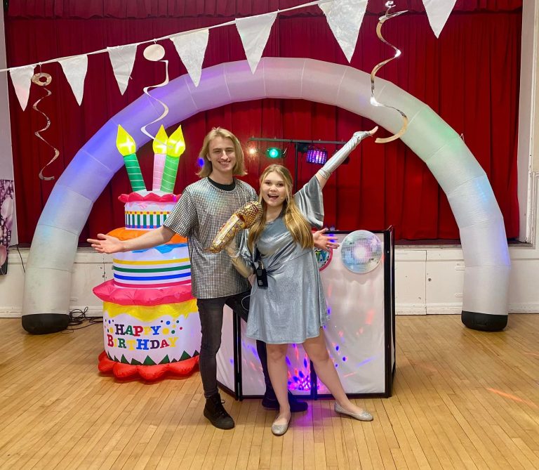 Two entertainers under a light-up arch next to a giant birthday cake inflatable