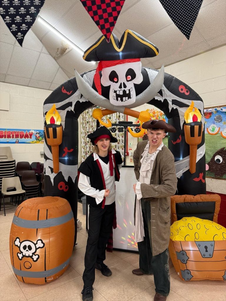 Two pirate entertainers beside a pirate ship arch with barrel and treasure chest inflatables