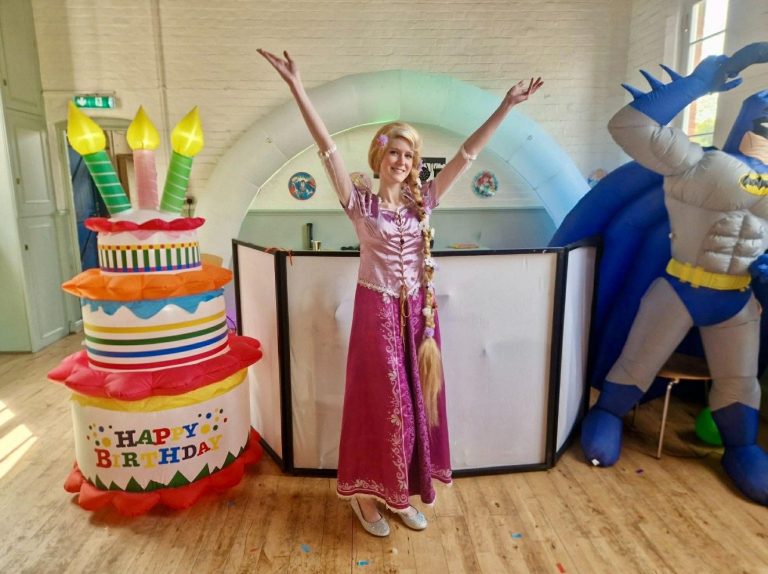 Rapunzel entertainer with arms raised next to a giant cake inflatable and Batman inflatable