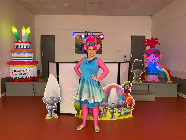 Poppy entertainer with pink hair beside Trolls decorations and a cake inflatable