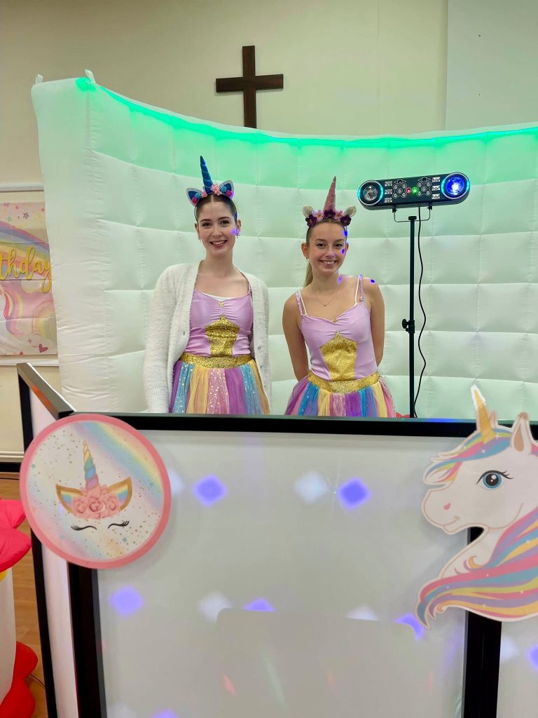Two unicorn entertainers with unicorn horns behind a decorated DJ booth