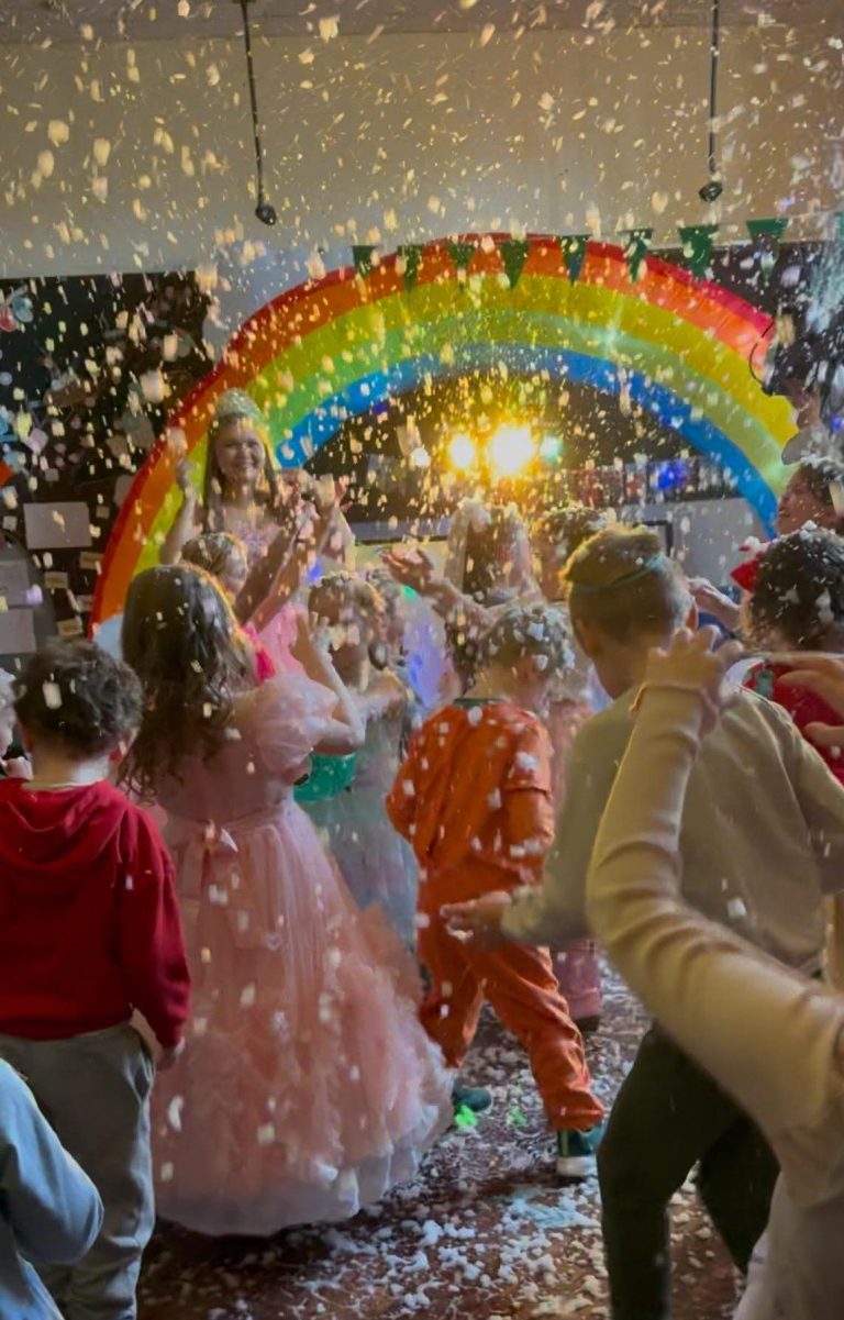 Glinda dancing under a rainbow arch as confetti falls on children